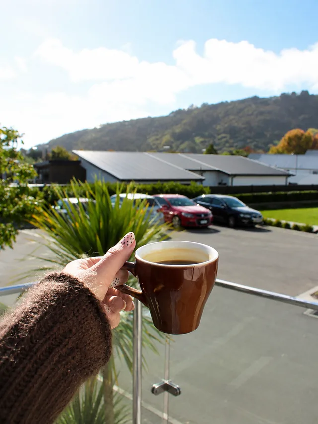 A hand holding a coffee outside Silos Cafe with Heathcote Valley behind it